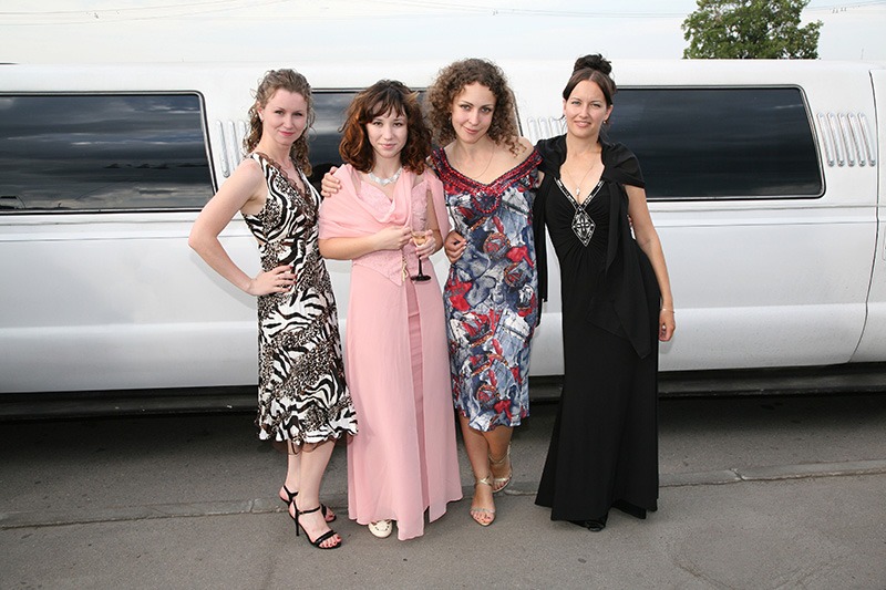 Four women in fancy dresses pose for a picture in front of a white limousine