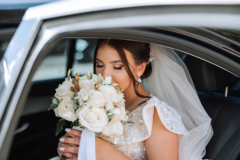 The bride is sitting in a black car with a bouquet of flowers in her hands and looks out of the car. Portrait of a rather shy bride in a car. Bride, smile, emotions. Weddings and Anniversaries